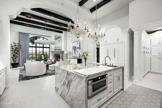 kitchen with a kitchen island with sink, a notable chandelier, light stone counters, beam ceiling, and white cabinets