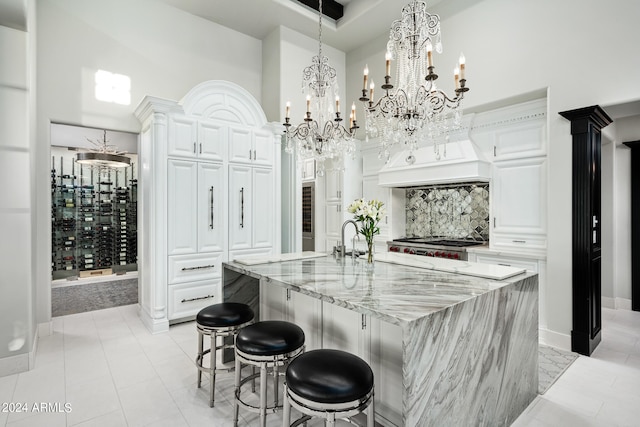 kitchen featuring a kitchen island with sink, a high ceiling, white cabinetry, a chandelier, and light stone counters