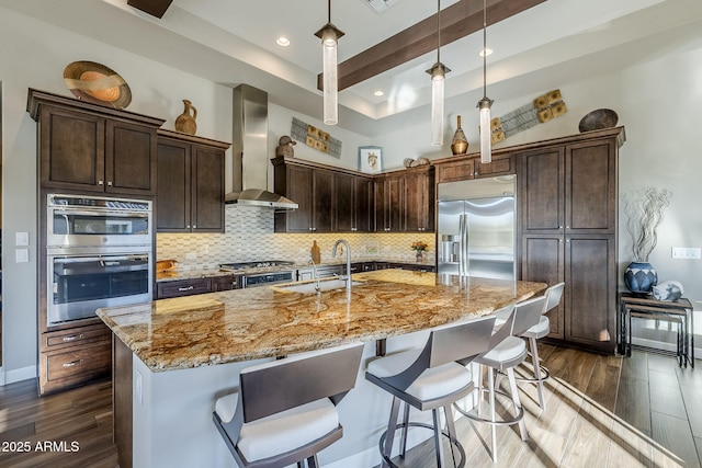 kitchen featuring a kitchen island with sink, wall chimney exhaust hood, stainless steel appliances, and decorative light fixtures