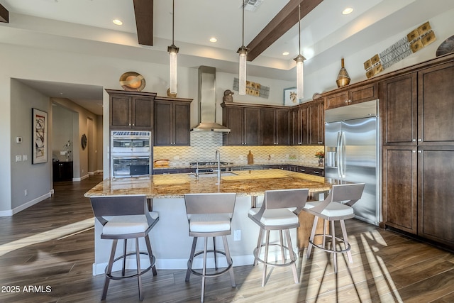 kitchen featuring light stone countertops, wall chimney exhaust hood, stainless steel appliances, a large island with sink, and decorative light fixtures