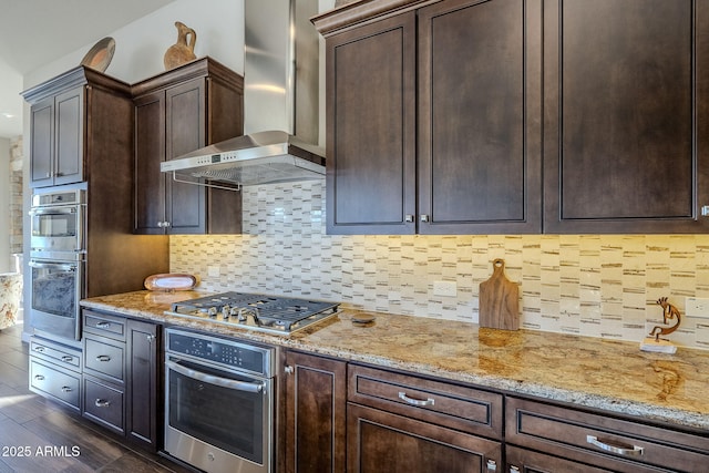 kitchen with dark brown cabinetry, light stone countertops, wall chimney range hood, decorative backsplash, and appliances with stainless steel finishes