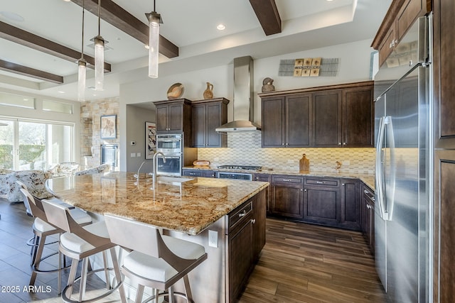 kitchen featuring wall chimney exhaust hood, an island with sink, decorative light fixtures, light stone counters, and stainless steel appliances