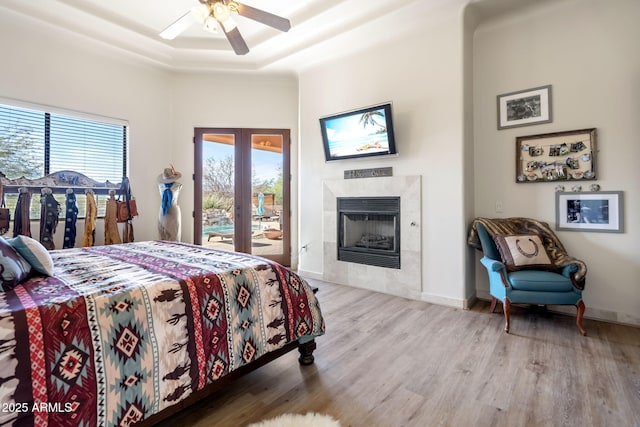 bedroom with wood-type flooring, access to outside, a raised ceiling, a tile fireplace, and ceiling fan