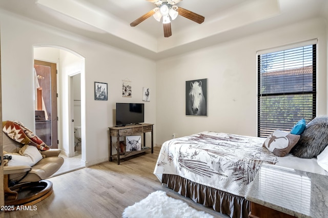 bedroom with ceiling fan, a tray ceiling, ensuite bath, and light wood-type flooring