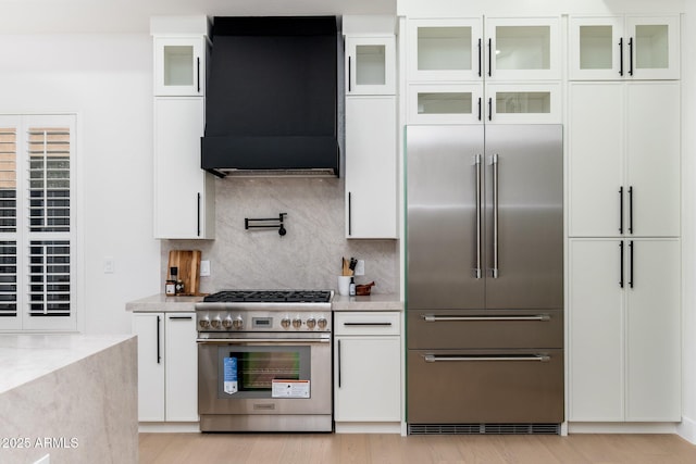 kitchen with premium appliances, exhaust hood, white cabinetry, tasteful backsplash, and light wood-style floors
