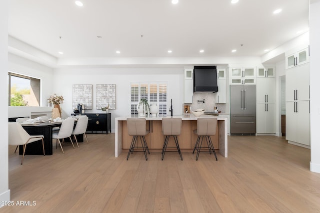 kitchen with stainless steel built in refrigerator, wall chimney range hood, light wood-style flooring, a large island with sink, and recessed lighting