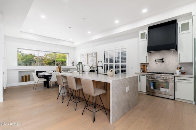kitchen featuring stainless steel range, wall chimney exhaust hood, a raised ceiling, light wood-style floors, and recessed lighting