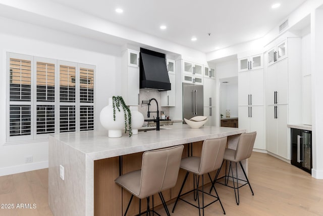kitchen with custom range hood, wine cooler, light wood-style floors, high end refrigerator, and a breakfast bar