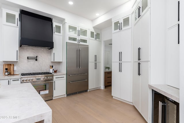 kitchen with premium appliances, range hood, beverage cooler, light countertops, and light wood-type flooring