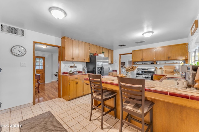 kitchen with tile countertops, visible vents, stainless steel range with gas stovetop, fridge with ice dispenser, and a peninsula