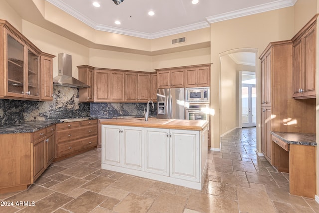 kitchen with stainless steel appliances, wall chimney range hood, butcher block countertops, crown molding, and a center island with sink