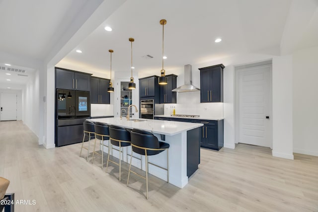 kitchen featuring wall chimney exhaust hood, light hardwood / wood-style floors, hanging light fixtures, a breakfast bar area, and an island with sink