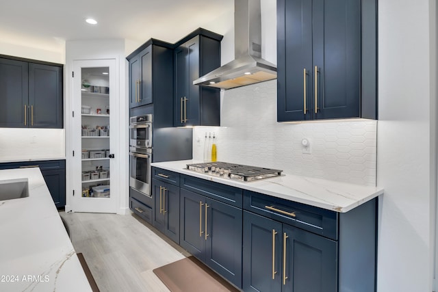 kitchen featuring blue cabinets, wall chimney exhaust hood, decorative backsplash, light hardwood / wood-style floors, and stainless steel appliances