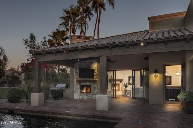 patio terrace at dusk with ceiling fan, exterior kitchen, and an outdoor stone fireplace