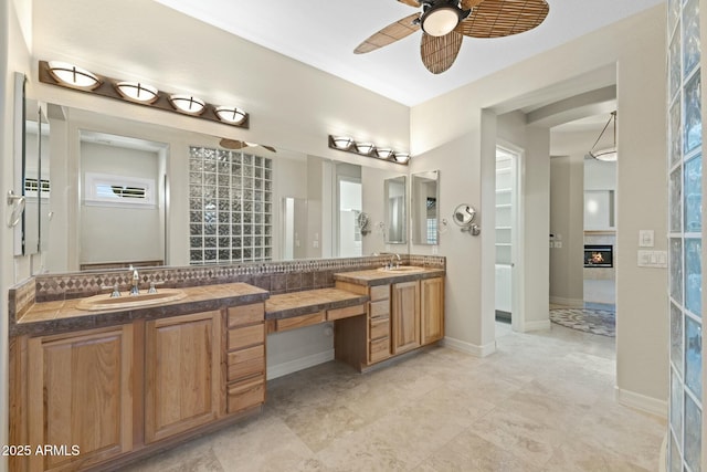 bathroom with a ceiling fan, a glass covered fireplace, a sink, and double vanity