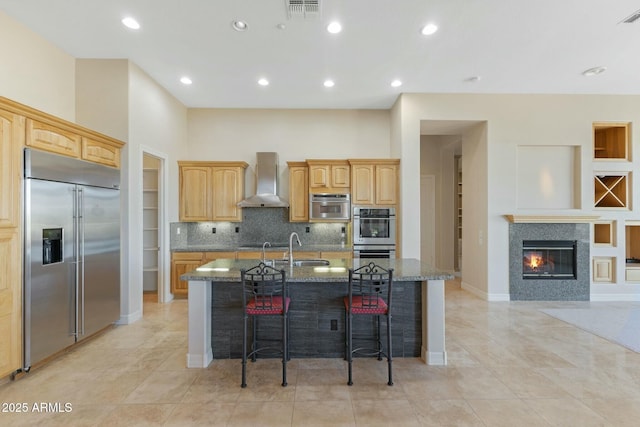 kitchen with a center island with sink, stainless steel appliances, a sink, wall chimney range hood, and dark stone counters