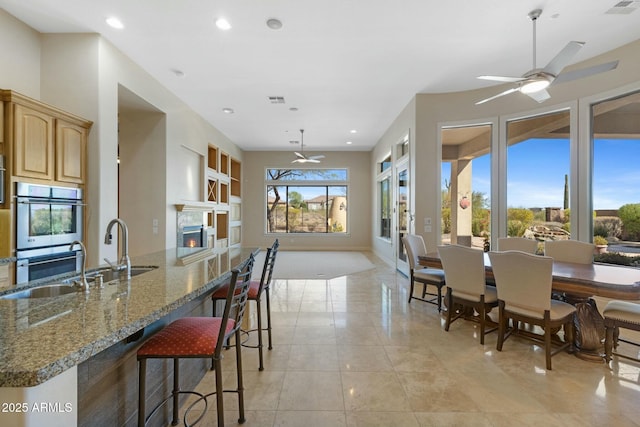 kitchen with stone countertops, a ceiling fan, a lit fireplace, stainless steel double oven, and a kitchen bar