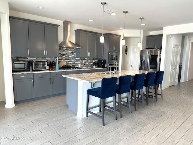 kitchen featuring light stone counters, wall chimney exhaust hood, an island with sink, hanging light fixtures, and appliances with stainless steel finishes