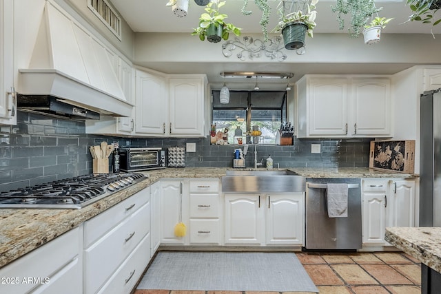 kitchen with sink, custom exhaust hood, white cabinetry, light stone counters, and appliances with stainless steel finishes