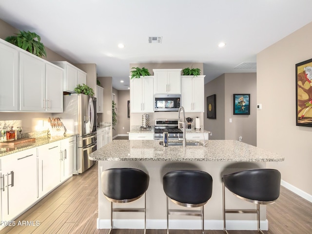 kitchen featuring a center island with sink, appliances with stainless steel finishes, light wood-type flooring, white cabinets, and light stone counters