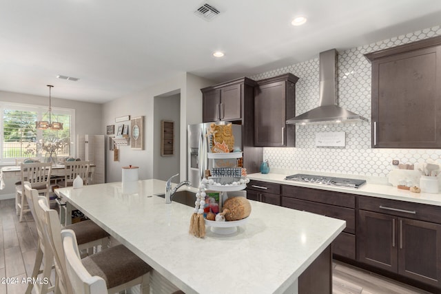 kitchen featuring a kitchen island with sink, wall chimney exhaust hood, pendant lighting, light wood-type flooring, and appliances with stainless steel finishes