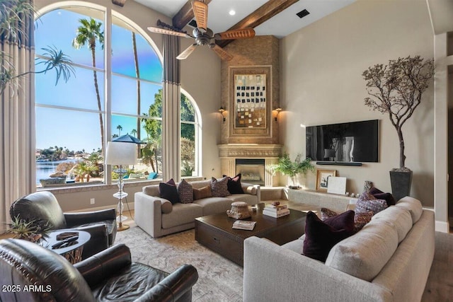 living room with ceiling fan, a water view, a fireplace, beamed ceiling, and light wood-type flooring