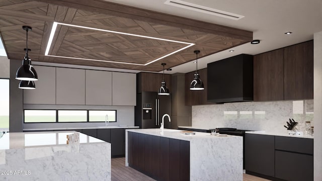 kitchen with a center island with sink, refrigerator with ice dispenser, backsplash, wall chimney range hood, and pendant lighting