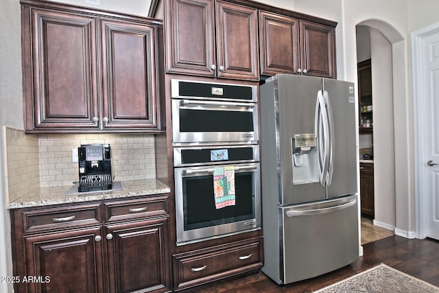 kitchen with dark wood-type flooring, stainless steel appliances, dark brown cabinetry, light stone countertops, and decorative backsplash