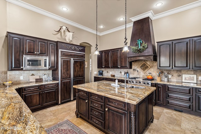 kitchen featuring premium range hood, built in appliances, dark brown cabinetry, light stone countertops, and decorative light fixtures