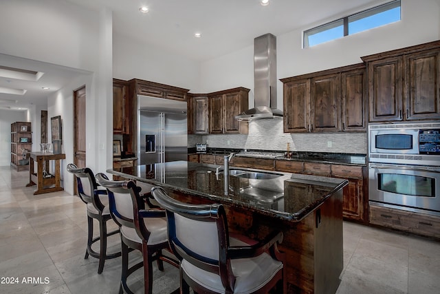 kitchen with a kitchen island with sink, wall chimney range hood, built in appliances, a towering ceiling, and tasteful backsplash