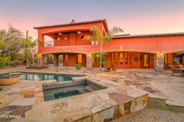 back house at dusk featuring a pool with hot tub, a patio area, and a balcony