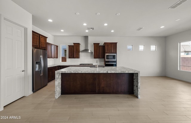 kitchen featuring appliances with stainless steel finishes, tasteful backsplash, light stone counters, wall chimney range hood, and a center island with sink