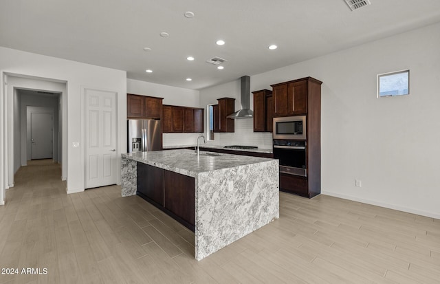 kitchen with sink, wall chimney range hood, backsplash, an island with sink, and appliances with stainless steel finishes