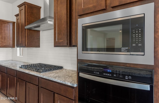 kitchen featuring tasteful backsplash, light stone counters, wall chimney range hood, and appliances with stainless steel finishes