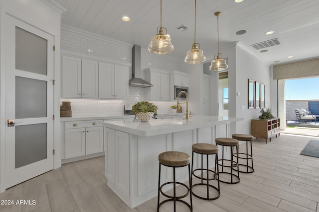 kitchen featuring pendant lighting, wall chimney range hood, white cabinets, and a kitchen island with sink