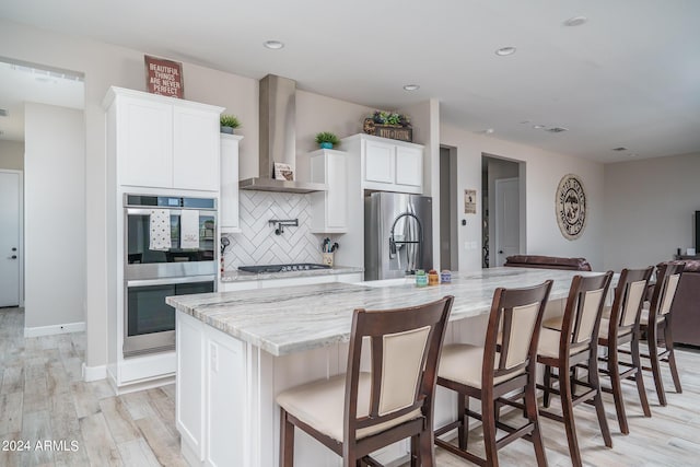 kitchen with appliances with stainless steel finishes, white cabinets, a kitchen breakfast bar, wall chimney range hood, and a spacious island