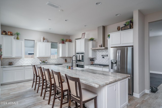 kitchen with wall chimney exhaust hood, white cabinetry, stainless steel appliances, and a kitchen island with sink