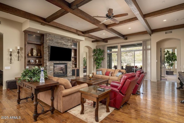 living room featuring ceiling fan, beam ceiling, coffered ceiling, light hardwood / wood-style floors, and a stone fireplace