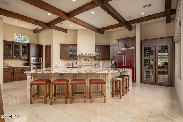 kitchen featuring a breakfast bar area, a large island with sink, built in appliances, light stone countertops, and wall chimney exhaust hood