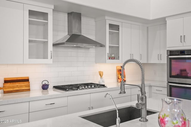 7775 E Fledgling Dr, Scottsdale, AZ 85255 - kitchen with wall chimney range hood, stainless steel appliances, white cabinetry, glass insert cabinets, and decorative backsplash