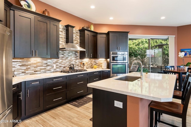 kitchen featuring sink, wall chimney range hood, an island with sink, a breakfast bar, and appliances with stainless steel finishes