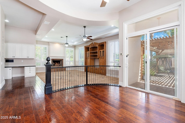 unfurnished living room with a ceiling fan, wood-type flooring, a tray ceiling, baseboards, and a warm lit fireplace