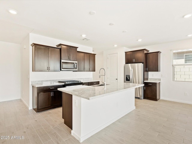 kitchen with light wood-type flooring, a center island with sink, a sink, stainless steel appliances, and dark brown cabinetry