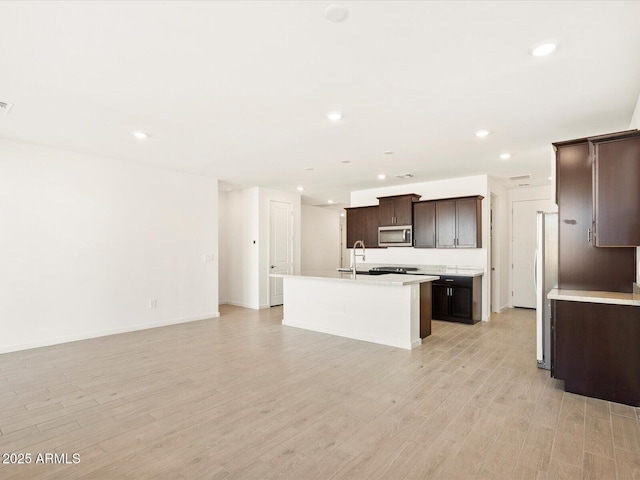 kitchen with a center island with sink, light countertops, dark brown cabinetry, stainless steel microwave, and light wood-type flooring