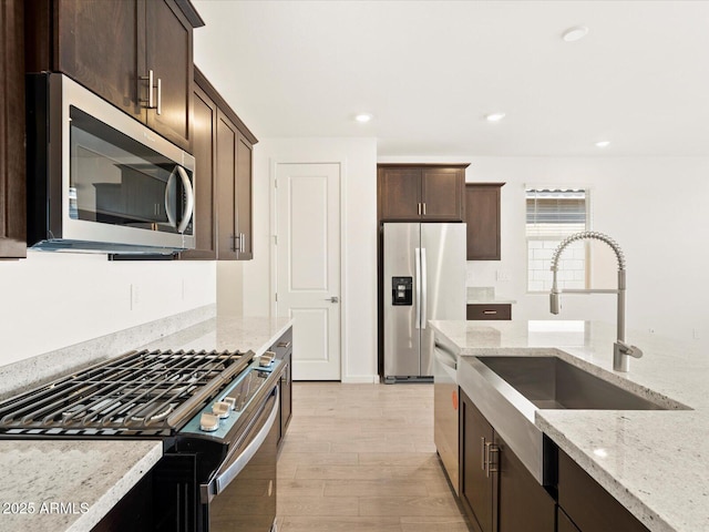 kitchen featuring light stone countertops, a sink, stainless steel appliances, dark brown cabinetry, and light wood-type flooring