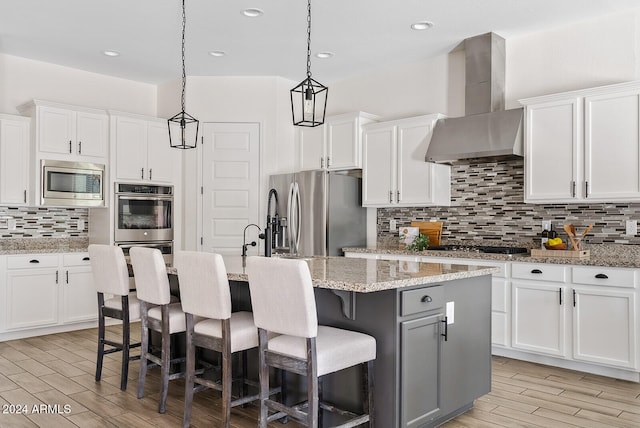 kitchen featuring an island with sink, light stone countertops, stainless steel appliances, white cabinets, and wall chimney range hood