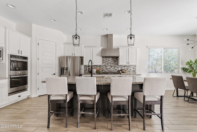 kitchen featuring stainless steel appliances, light stone countertops, an island with sink, and wall chimney range hood