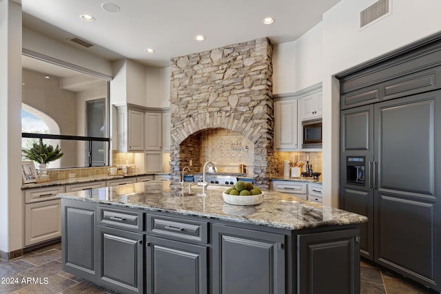 kitchen with backsplash, built in appliances, gray cabinets, light stone countertops, and an island with sink