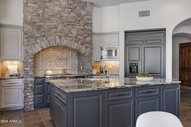 kitchen featuring tasteful backsplash, sink, built in appliances, stone countertops, and an island with sink