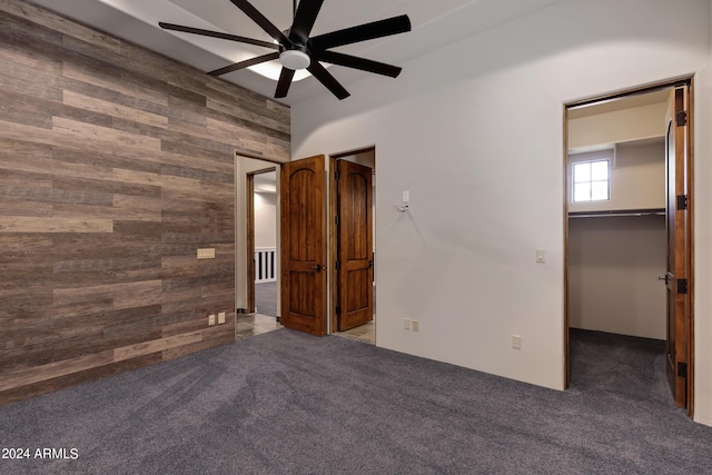 unfurnished bedroom with wood walls, dark carpet, a walk in closet, ceiling fan, and a closet